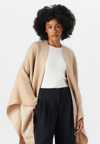 Beige ullsjal med åpen front og sidefringe, båret over en hvit ribbestrikket topp og mørke skreddersydde bukser. Glatt tekstur, enkelt design.
