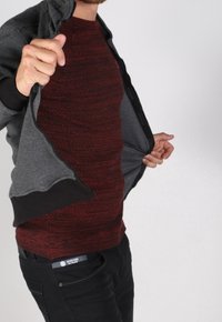 Chaqueta gris con un ribete negro sólido, llevada sobre un suéter rojo texturizado. Jeans negros visibles en la cintura, mostrando un estilo casual en capas.