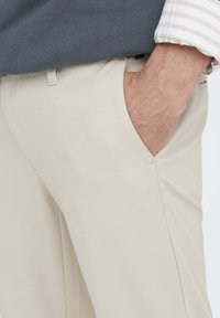 Braccio maschile con mano nella tasca dei pantaloni beige, indossando una maglietta grigia scura e una camicia bianca a maniche lunghe con riflessi chiari.