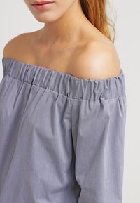 Off-shoulder blus i lätt ljusblått tyg med en rynkad elastisk halsringning. Har en slät yta och ett subtilt rutig mönster.