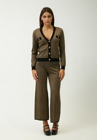 Bruine metalen gebreide cardigan met een diepe V-hals, zwarte afwerking en gouden knopen, gecombineerd met bijpassende wijde broek en zwarte hoge hakken.