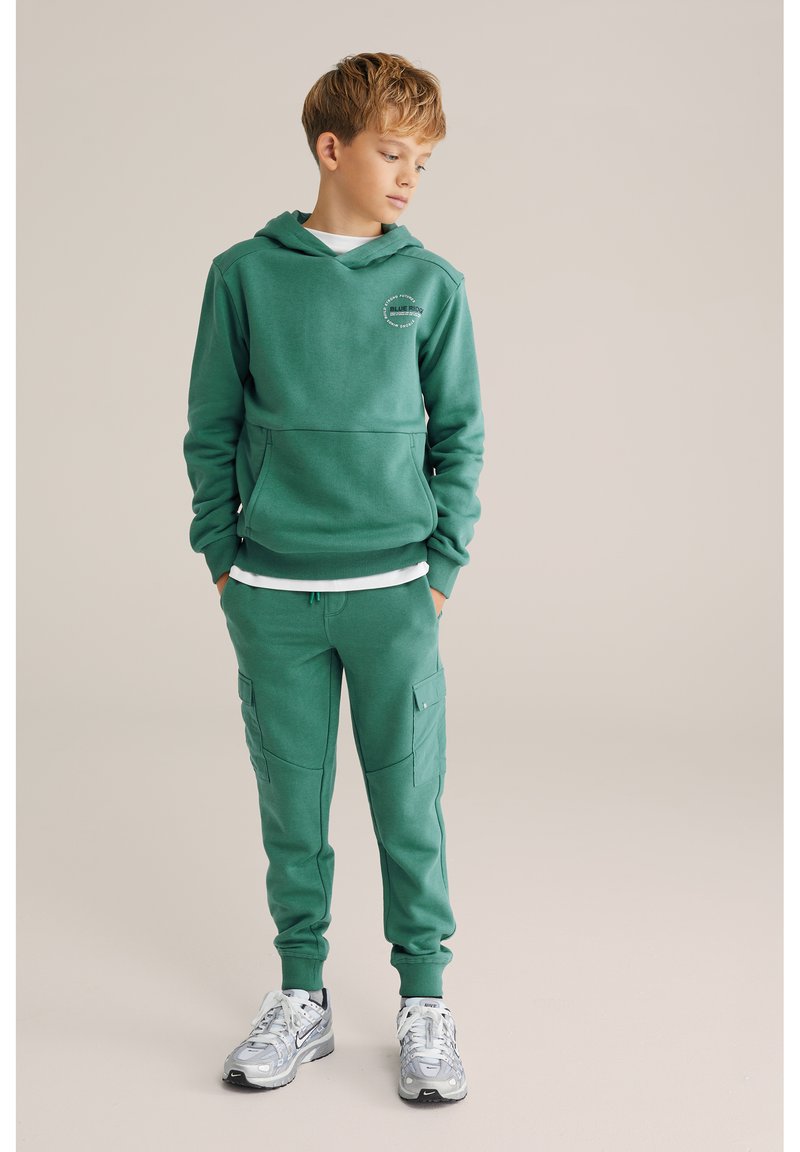 Roheline kapuutsiga dressipluus eesmise tasku ja logoga, koos sobivate küljetaskute ja kaubafailidega dressipükstega. Mudel kannab hõbedaseid spordijalatseid.