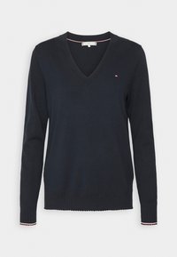 Pull à col en V bleu marine à manches longues avec poignets et ourlet côtelés, arborant un petit logo brodé rouge, blanc et bleu sur la poitrine gauche.