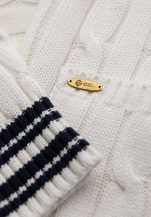 Maglione bianco a trecce con un motivo testurizzato. Presenta polsini a righe blu navy e bianchi e una piccola etichetta dorata con la scritta "OMBRE HERITAGE."