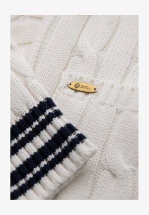Maglione bianco a trecce con un motivo testurizzato. Presenta polsini a righe blu navy e bianchi e una piccola etichetta dorata con la scritta "OMBRE HERITAGE."