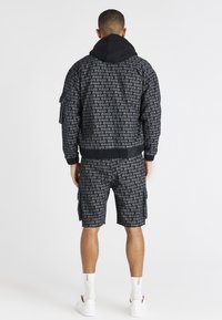 Bomber nero con motivo di testo ripetuto, abbinato a pantaloncini cargo neri. Presenta un cappuccio e polsini elastici, con calzini bianchi e sneakers.