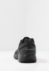 Zapatilla deportiva negra con parte superior texturizada, suela de goma y cuello acolchado. Presenta el logo "Reebok" en gris en el talón.