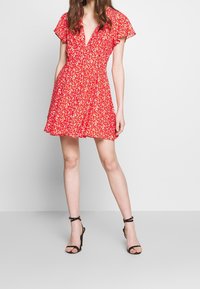 Vestido floral rojo con mangas cortas acampanadas, escote en V, cintura ceñida y falda acampanada; combinado con sandalias de tacón alto negras con tiras.