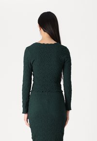 Long-sleeve, dark green top with a textured fabric. Features a scalloped neckline and hem, fitted design, and matching skirt.