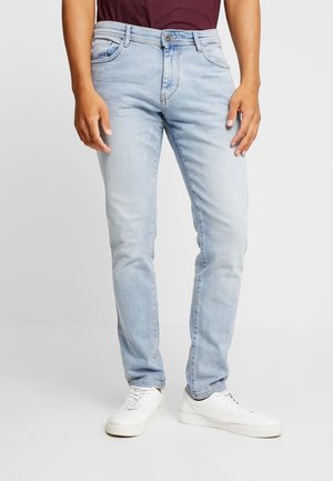 Mężczyzna ubrany w jasnoniebieskie dopasowane jeansy, białe sneakersy i bordową koszulę, stojący w luźnej pozie na jednolitym tle.