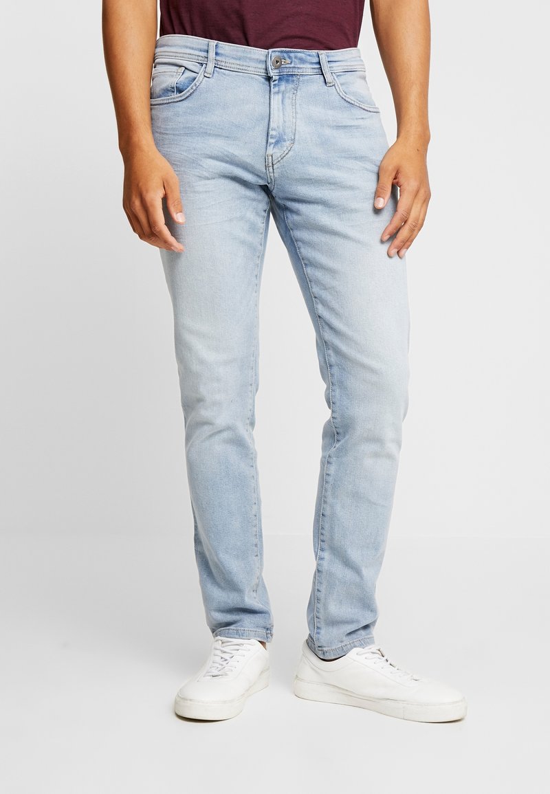 Homme portant un jean slim bleu clair, des baskets blanches et une chemise bordeaux, debout avec une posture détendue sur un fond uni.