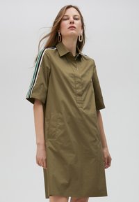 Olive green dress with a collared neck, short sleeves, and two side pockets. Features contrasting stripes on sleeves and a button-less placket.