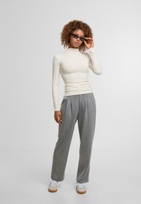 Langärmliges, figurbetontes cremefarbenes Oberteil, kombiniert mit lockeren grauen Hosen, die aus weichem Stoff gefertigt sind und seitliche Falten aufweisen; das Model trägt dunkle Sonnenbrillen.