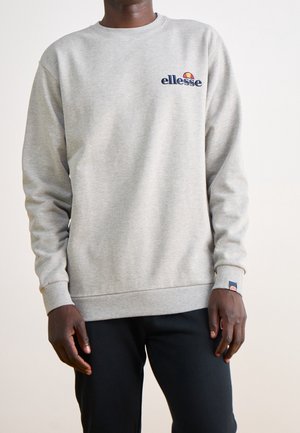 Homme portant un sweat-shirt à col rond gris clair ellesse avec un logo sur la poitrine et le poignet, associé à un pantalon noir, debout devant un fond blanc.