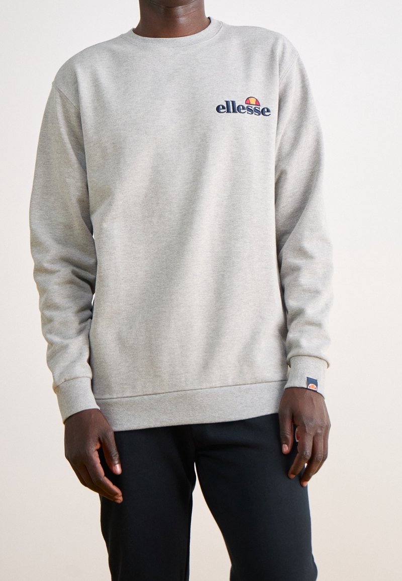 Homme portant un sweat-shirt à col rond gris clair ellesse avec un logo sur la poitrine et le poignet, associé à un pantalon noir, debout devant un fond blanc.