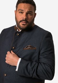 Donkerblauwe textuur jas met bruine leren accenten en knopen. Inclusief een geruite pochet. Voorkant, model houdt de jas vast.