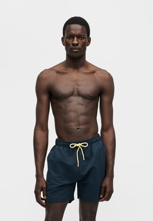 Fit černoch stojaci, oblečený v tmavomodré plavky s béžovým šnúrkou, na jednoduchom bielom pozadí.