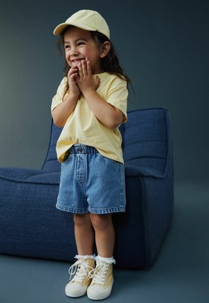 Fată tânără cu șapcă și tricou galben, pantaloni scurți din denim și adidași galbeni, stând zâmbitoare cu mâinile aproape de față lângă un scaun albastru cu pernă.