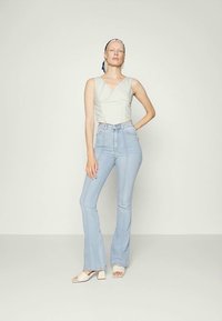 Mulher usando uma blusa sem mangas bege clara, jeans claros de cintura alta, sandálias brancas de salto, e um lenço padrão na cabeça, de pé contra um fundo liso.