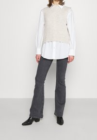 Beige pletený svetřík bez rukávů přes bílou košili, spárovaný se šedými zvonovými džíny a černými špičatými kotníkovými botami. Jednoduchý design.