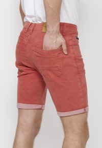 Pantalones cortos de mezclilla roja con dobladillo enrollado, que cuentan con bolsillos traseros y detalles de costura sutiles. El material tiene una textura desgastada para un aspecto casual.