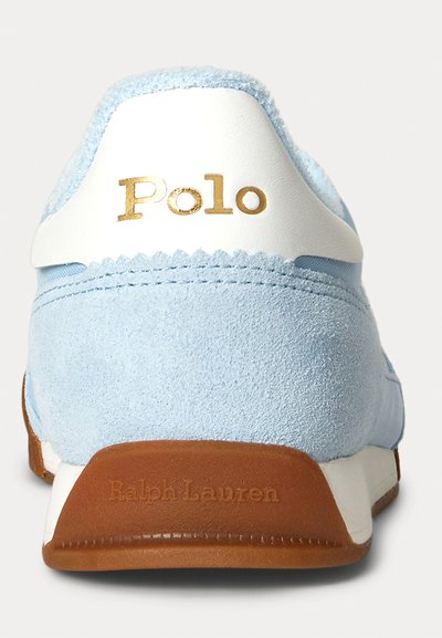 Sapatilha de camurça azul clara com um contra-forte branco e marcação em dourado "Polo". Apresenta uma sola de borracha castanha com uma discreta estampagem "Ralph Lauren".
