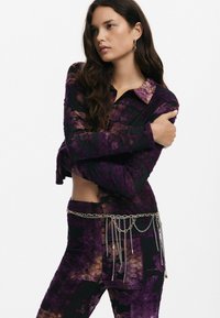 Camicetta corta a maniche lunghe con motivo viola e pantaloni abbinati. Caratterizzata da un tessuto testurizzato e una cintura in catena argentata con dettagli pendenti.