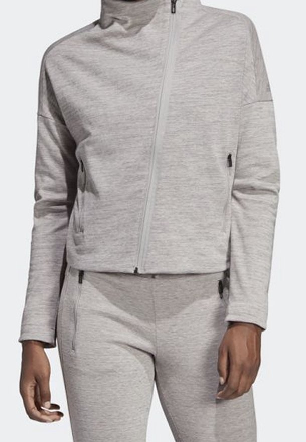 Femme portant une veste zippée gris clair et un pantalon de survêtement ajusté assorti, tous deux avec des poches zippées, sur un fond uni.