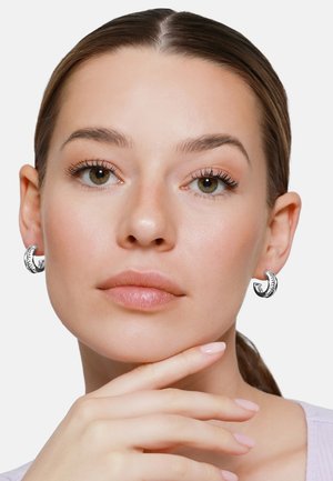 Giovane donna con capelli tirati indietro che indossa orecchini a cerchio d'argento, poggiando il mento sulla mano contro uno sfondo chiaro e uniforme.