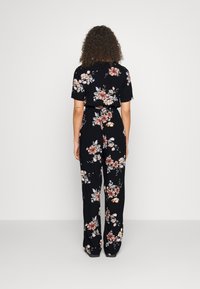 Kobieta z kręconymi włosami, ubrana w czarny, kwiecisty kombinezon z krótkim rękawem, stoi twarzą do gładkiej białej ściany.