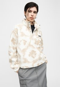 Νεαρό άτομο με κοντά σκούρα μαλλιά, φοράει μπεζ και λευκό φλις πουλόβερ με σχέδια και γκρι φούστα cargo, χέρια στις τσέπες, ουδέτερο φόντο.