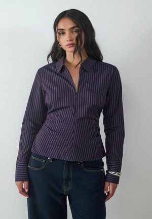 Navyblaues Hemd mit vertikalen Streifen in Lila, taillierte Silhouette, Kragen und lange Ärmel. Kombiniert mit dunkler Jeans und einem silbernen Armband.