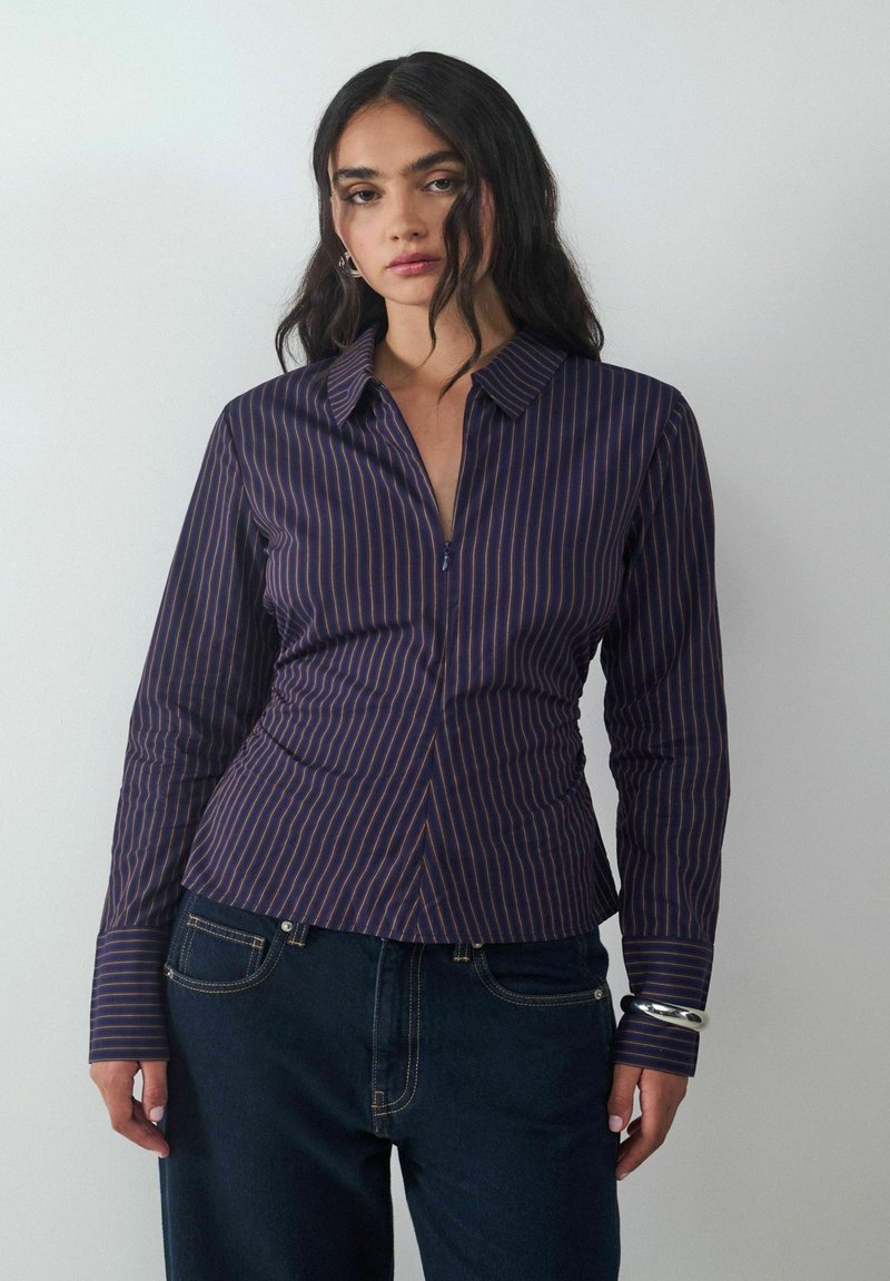 Navyblaues Hemd mit vertikalen Streifen in Lila, taillierte Silhouette, Kragen und lange Ärmel. Kombiniert mit dunkler Jeans und einem silbernen Armband.