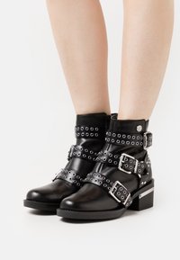 Bottines noires en cuir avec des sangles et des boucles à clous argentés, bout carré, petit talon bloc et accents décoratifs en argent sur le talon.