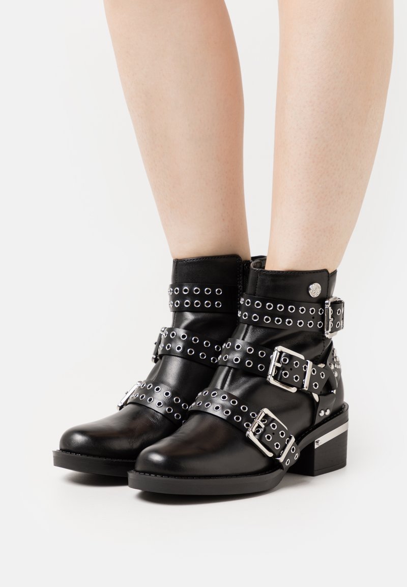 Bottines noires en cuir avec des sangles et des boucles à clous argentés, bout carré, petit talon bloc et accents décoratifs en argent sur le talon.