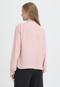 Light pink blouse with long sleeves, ruffled neckline, and gathered cuffs, paired with black pants. Smooth texture, relaxed fit.
