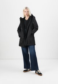 Black parka jacket with a fleece-lined hood, front zipper, and button closure. Paired with wide-leg jeans and black sneakers.