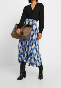 Black long-sleeve blouse with a V-neck, paired with a blue and black checkered pleated skirt. Model holds a brown leather tote bag.