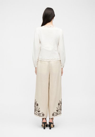 Γυναίκα με μακριά σκουρόχρωμα μαλλιά φοράει λευκό μπλουζάκι με μακριά μανίκια και μπεζ παντελόνια με φαρδιά πόδια και σκούρα κεντήματα με λουλούδια κοντά στους αστραγάλους, μαύρες γόβες.