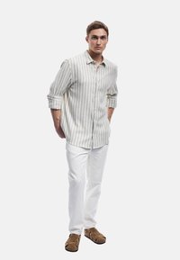 Camisa de botones a rayas color beige claro con mangas arremangadas, combinada con pantalones blancos y zapatos marrones con tiras, de pie sobre un fondo neutro.