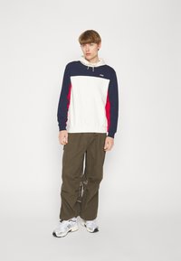 Felpa con cappuccio bianca, navy e rossa con logo; abbinata a pantaloni cargo verde oliva. Presenta un cappuccio con coulisse e polsini a costine.
