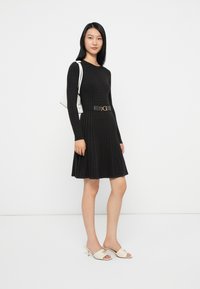 Abito lungo a maniche lunghe in maglia a coste nera con gonna svasata, vita cintrata da una cintura nera con accessori dorati. Borsa bianca e mule beige.
