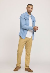 Chemise en denim bleu clair, superposée sur un t-shirt blanc. Un pantalon kaki et des chaussures marron complètent la tenue. Pose debout avec les mains ensemble.