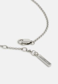 Ezüst lánc nyaklánc ovális szemekkel, homár kapoccsal és téglalap alakú címkével, amelyen az "Calvin Klein" felirat található. Simább, fémes felület.
