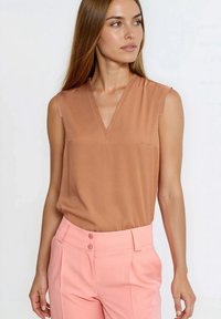 Beżowy top bez rękawów z dekoltem w serek i subtelną kieszonką na piersi, zestawiony z pastelowymi różowymi spodniami z wysokim stanem, zapinanymi na dwa guziki.