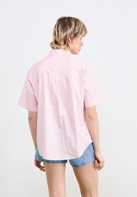 Camicia rosa chiaro a maniche corte in cotone, con una piega sul retro e un orlo curvo. Abbinata a pantaloni corti in denim chiaro, vista da dietro.