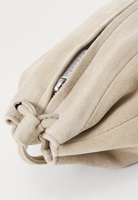 Béžová semišová taška s guľatým, zhromaždeným tvarom. Obsahuje strieborný zips, ktorý je čiastočne otvorený, čo odhaľuje hladký interiér a dekoratívne šnúrky.