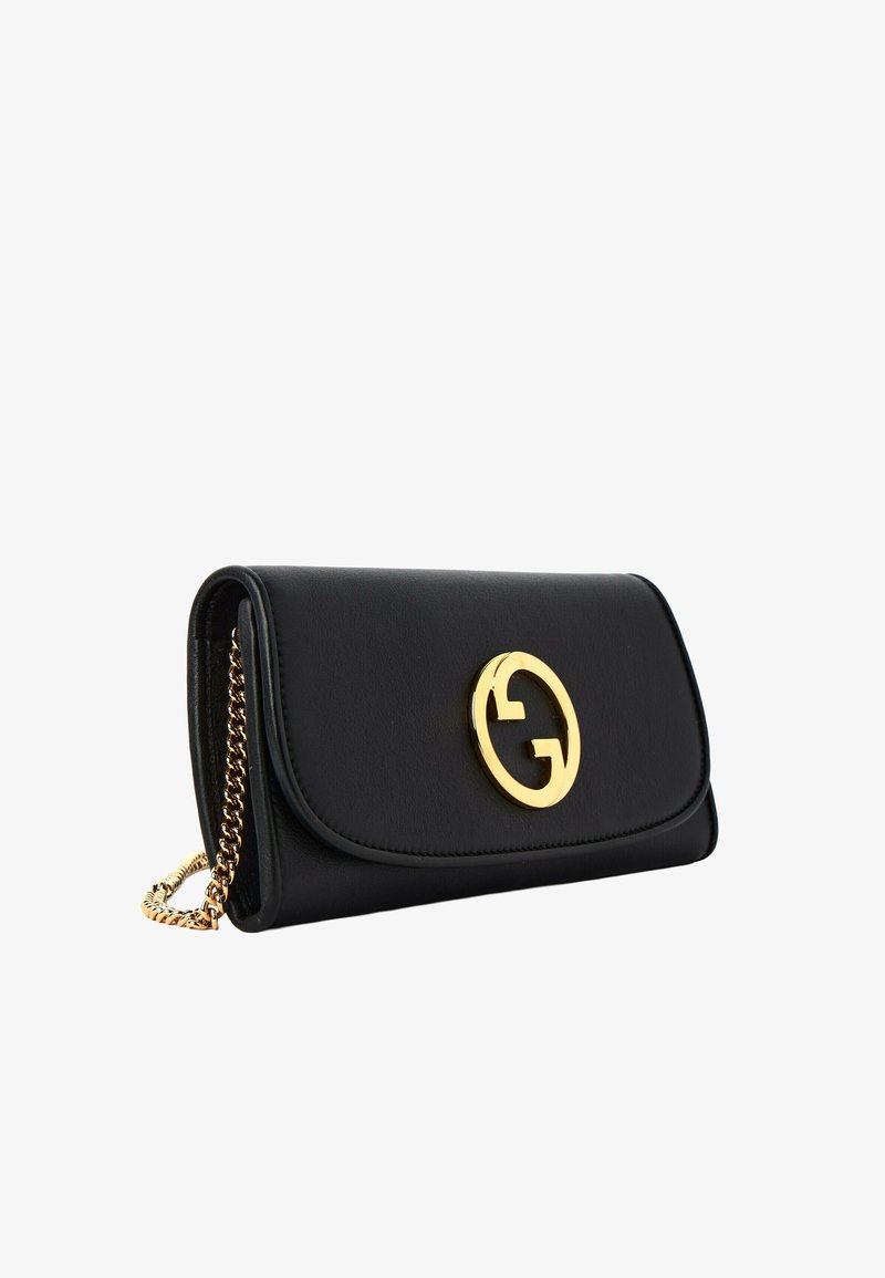 Pochette en cuir noir avec bandoulière chaîne dorée et emblème circulaire doré sur le rabat avant, présentée en angle sur fond blanc.