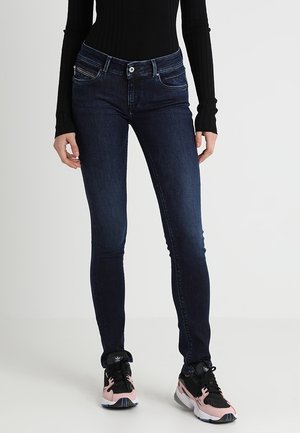 Wąskie ciemnoniebieskie jeansy noszone z czarną bluzką z długim rękawem oraz czarno-różowymi sneakersami na jednolitym jasnym tle.