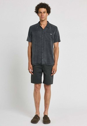 Homme debout portant une chemise noire à manches courtes délavée boutonnée, un short noir et des chaussures sombres sans lacets sur un fond uni.